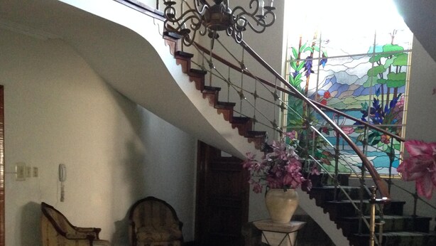 A spacious living area with a staircase leading to a balcony, a large window with a stained glass view, and a potted plant on a pedestal. - Hotel Regency (Lima)
