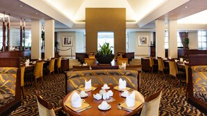 A hotel dining area with round tables set for four, each with white napkins, cutlery, and a centerpiece. The room features patterned carpet, wooden furniture, and a large chandelier.