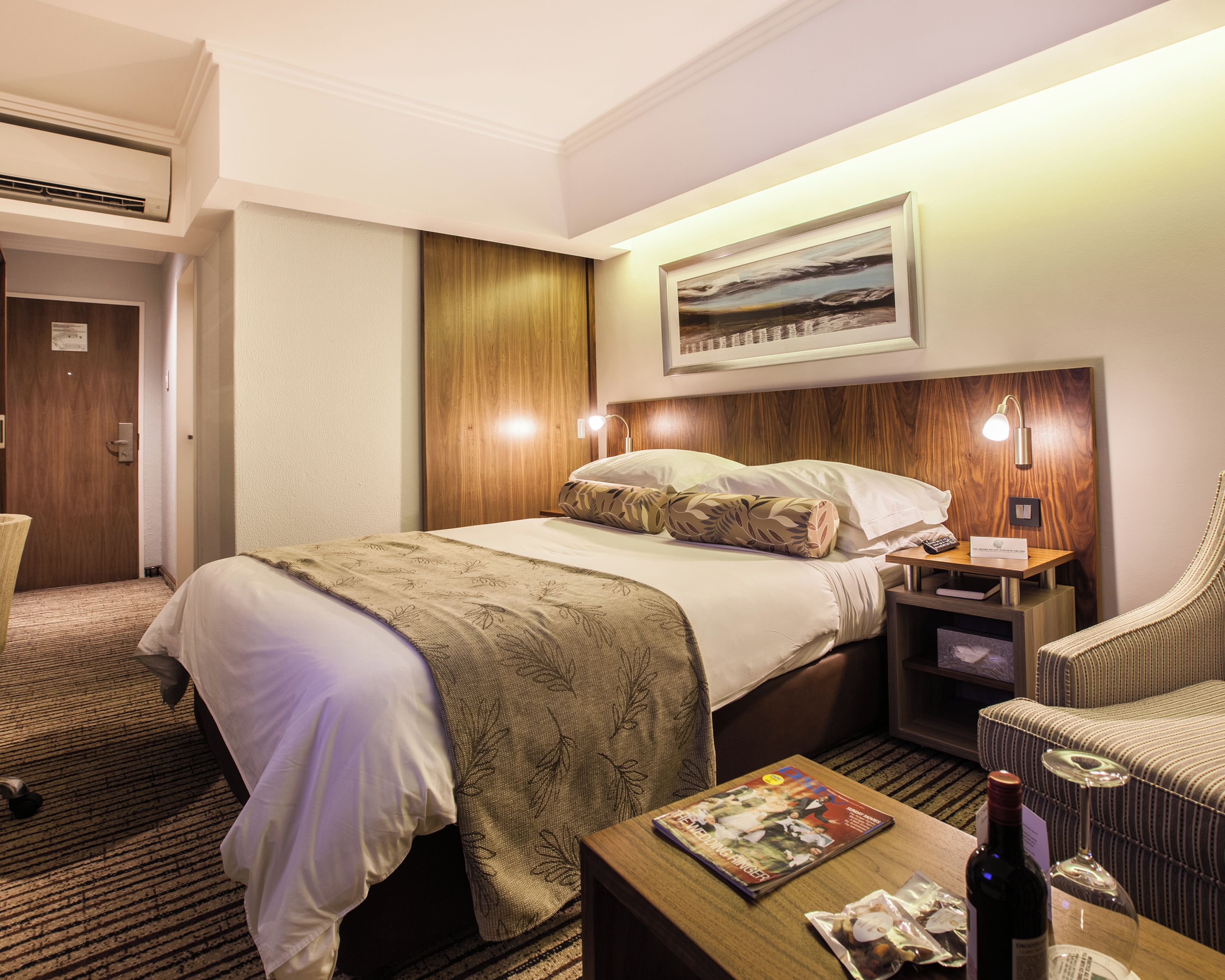 A hotel room with a large bed, a wooden headboard, a nightstand with a lamp, a seating area with a chair, and a framed picture on the wall.