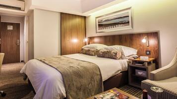A hotel room with a large bed, a wooden headboard, a nightstand with a lamp, a seating area with a chair, and a framed picture on the wall.