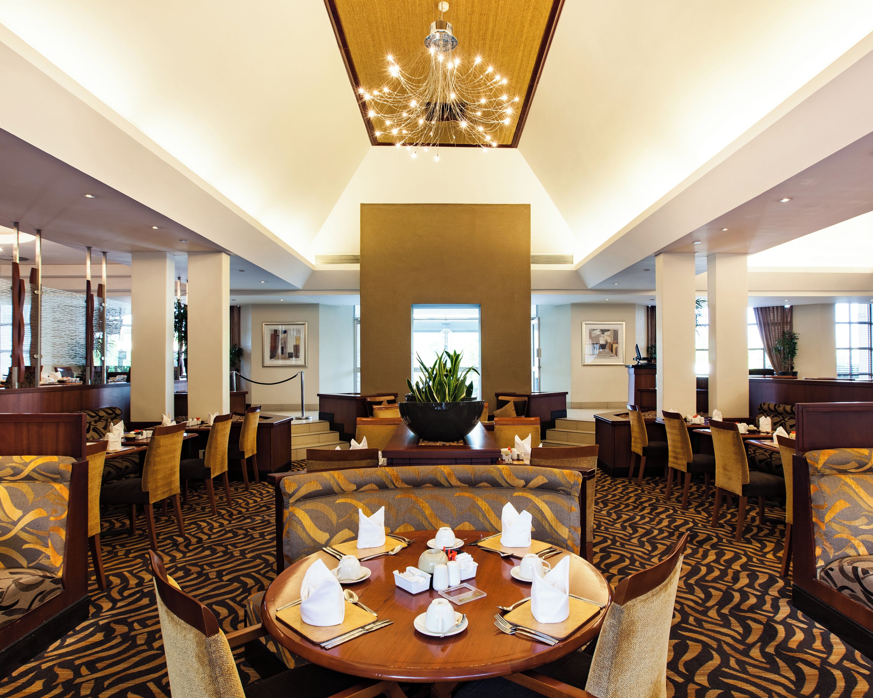 A hotel dining area with round tables set for four, each with white napkins, cutlery, and a centerpiece. The room features patterned carpet, wooden furniture, and a large chandelier.