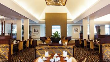 A hotel dining area with round tables set for four, each with white napkins, cutlery, and a centerpiece. The room features patterned carpet, wooden furniture, and a large chandelier.