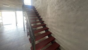Un escalier aux marches rouges et à la rampe métallique, menant à un espace lumineux offrant une vue sur l’extérieur.