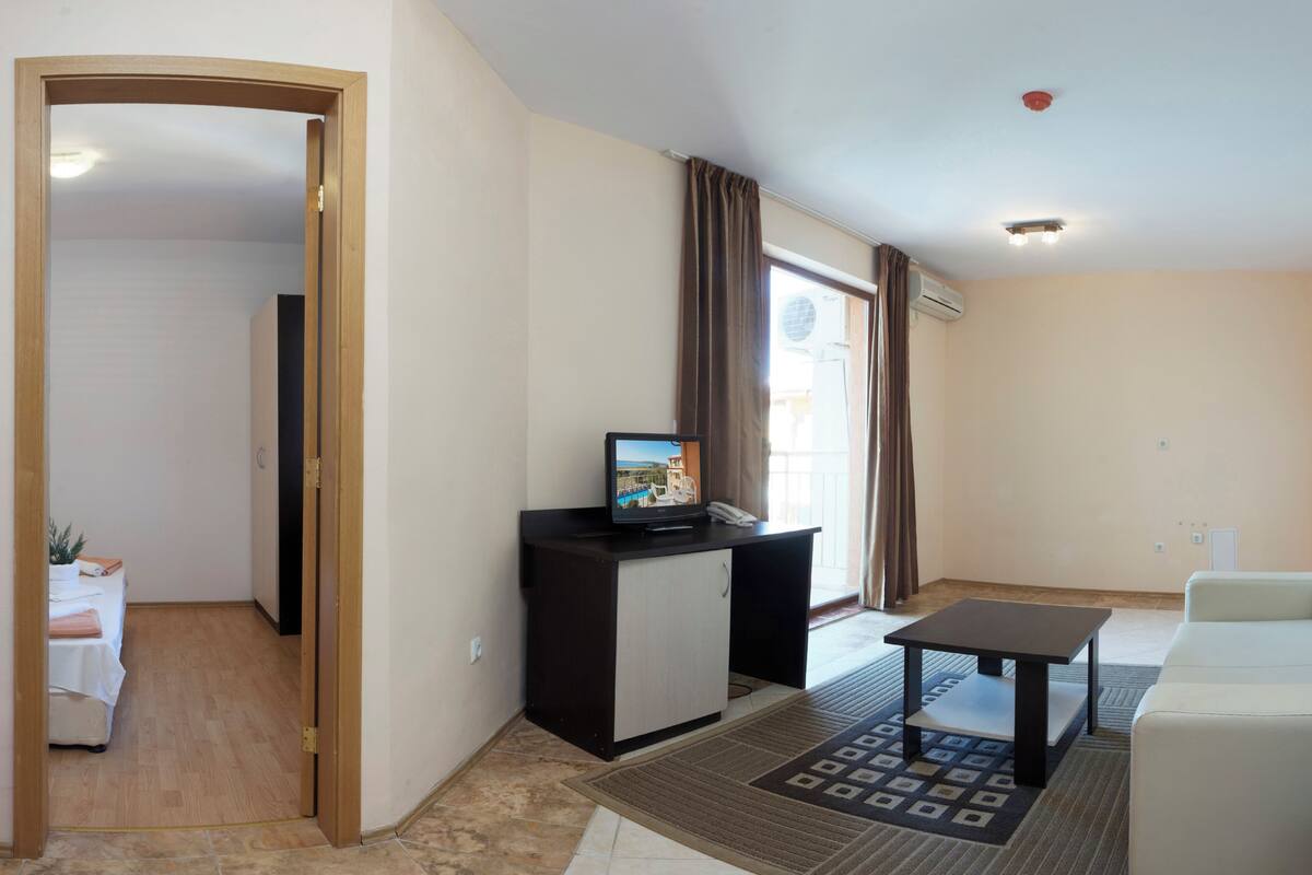 A living room with a sofa, a small table, a TV on a cabinet, a door leading to another room, and a window with curtains.