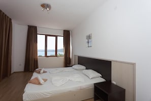 A bedroom with a large bed, a nightstand, a window with curtains, and a view of the sea.
