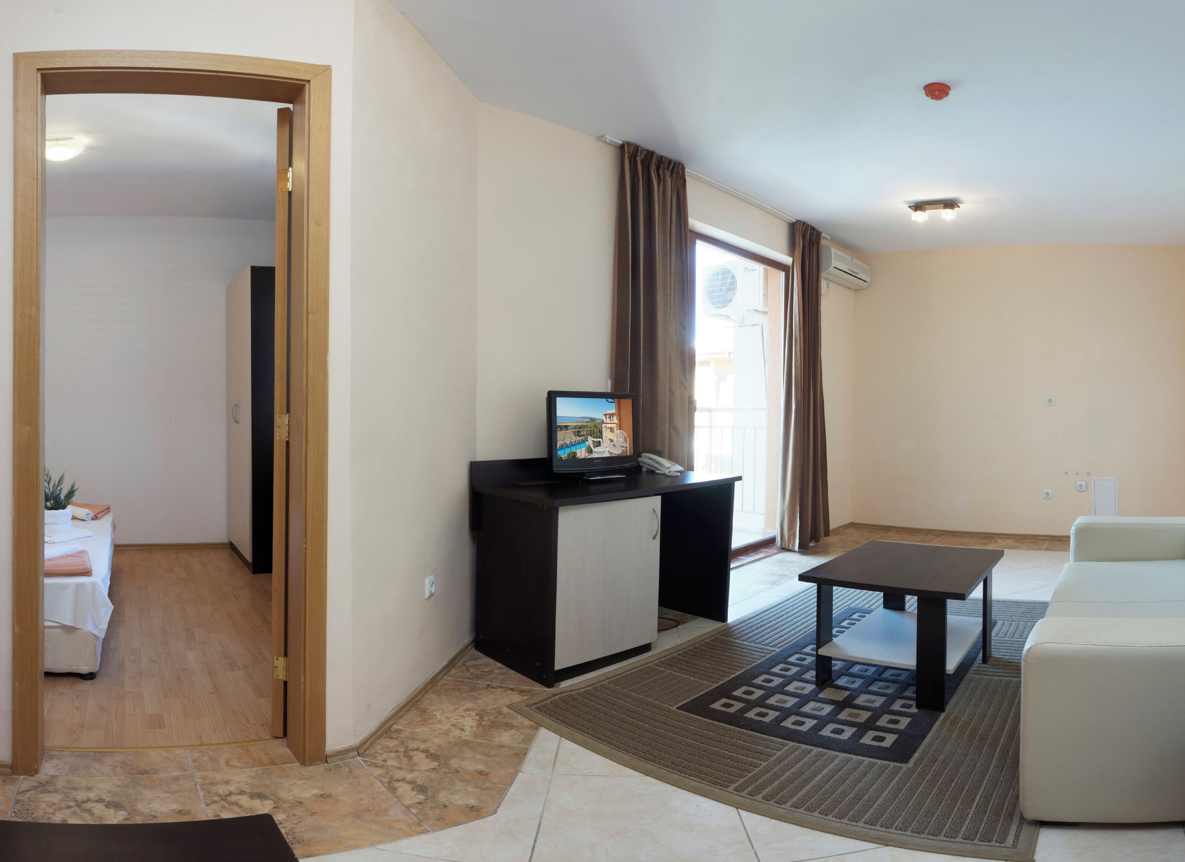 A living room with a sofa, a small table, a TV on a cabinet, a door leading to another room, and a window with curtains.