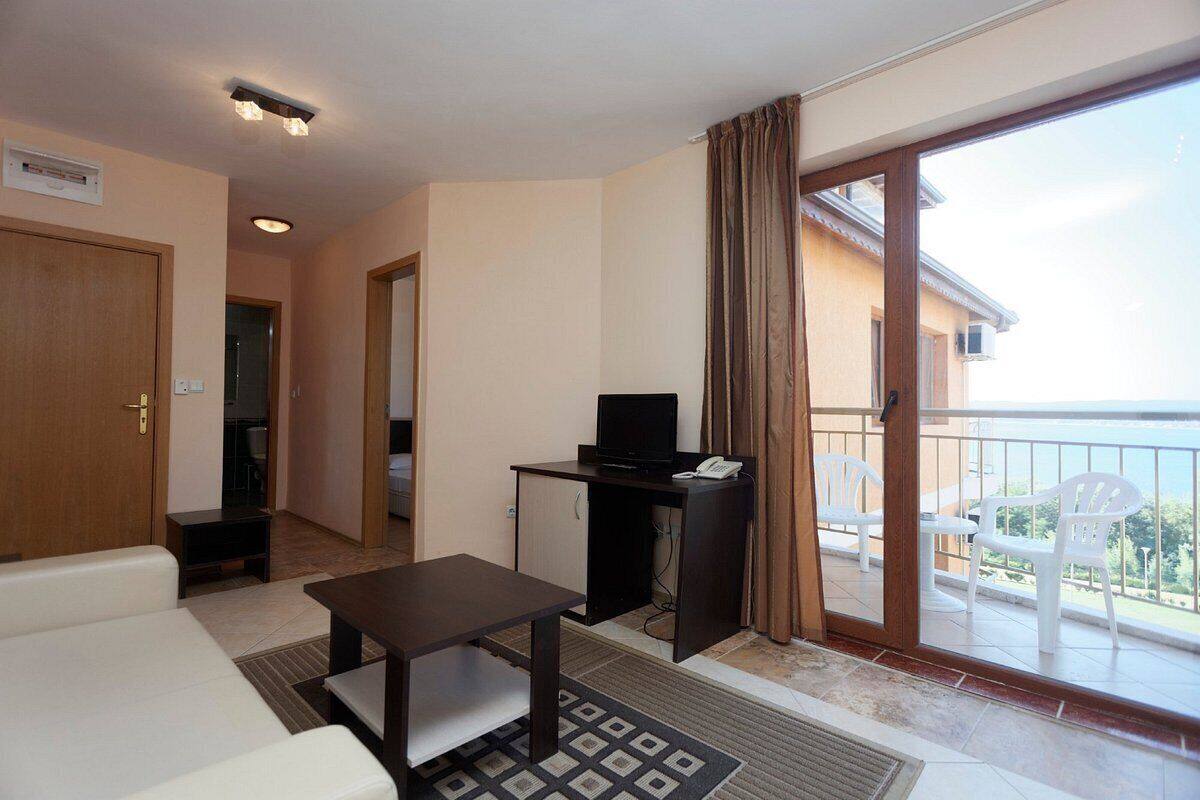 A room with a sofa, a small table, a TV on a cabinet, a sliding glass door to a balcony with chairs, and a door leading to another room.