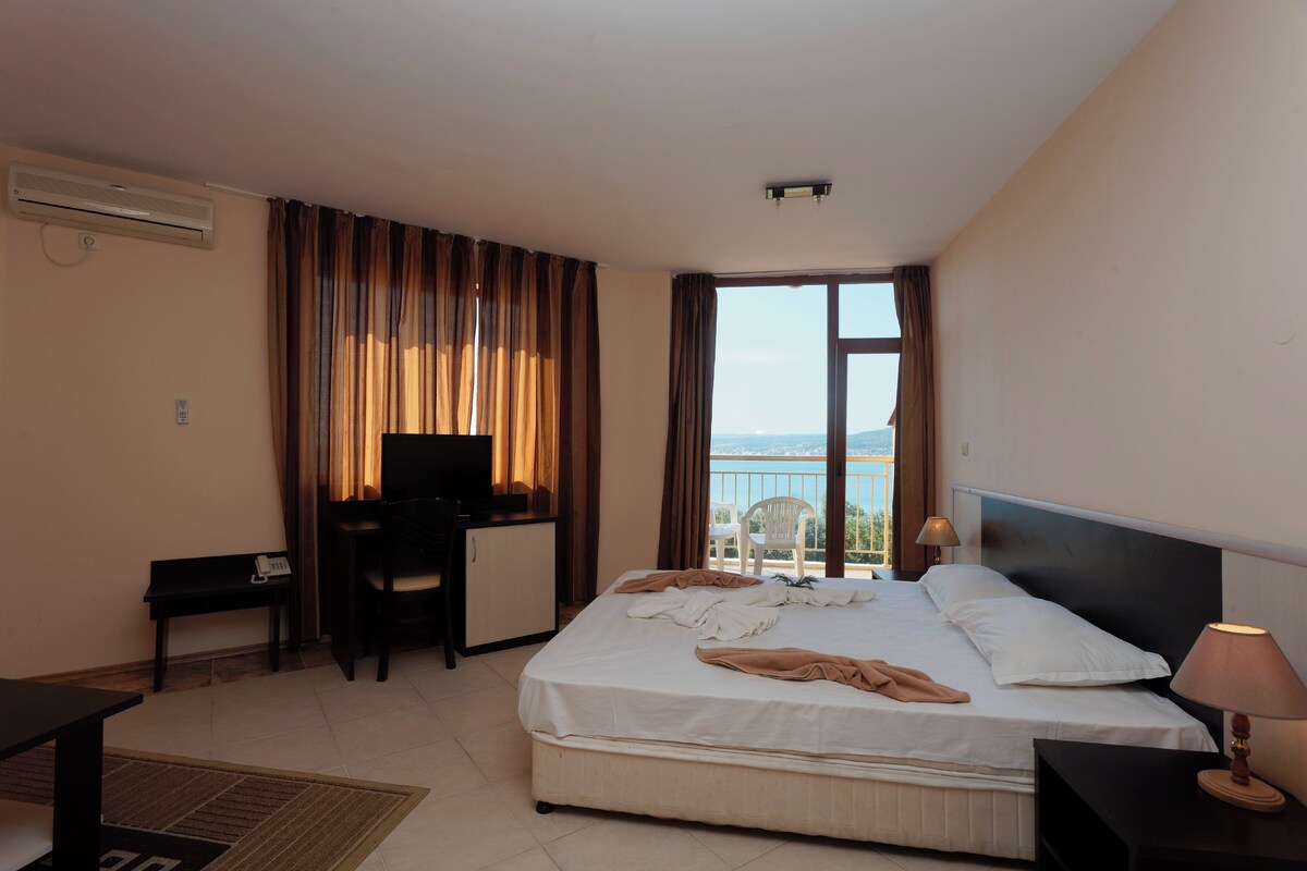 A hotel room with a bed, desk, chair, and a view of the sea.
