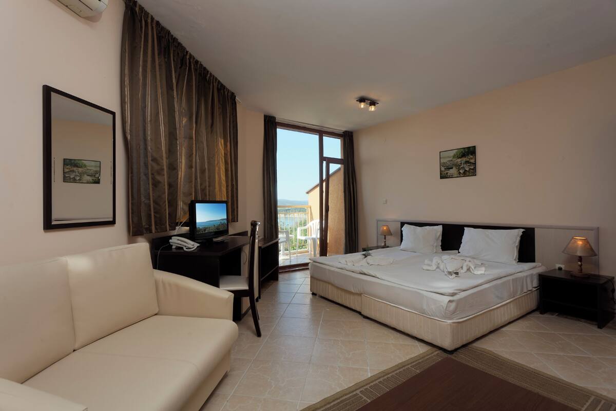 A hotel room with a bed, a desk with a computer, a couch, and a balcony with a view.