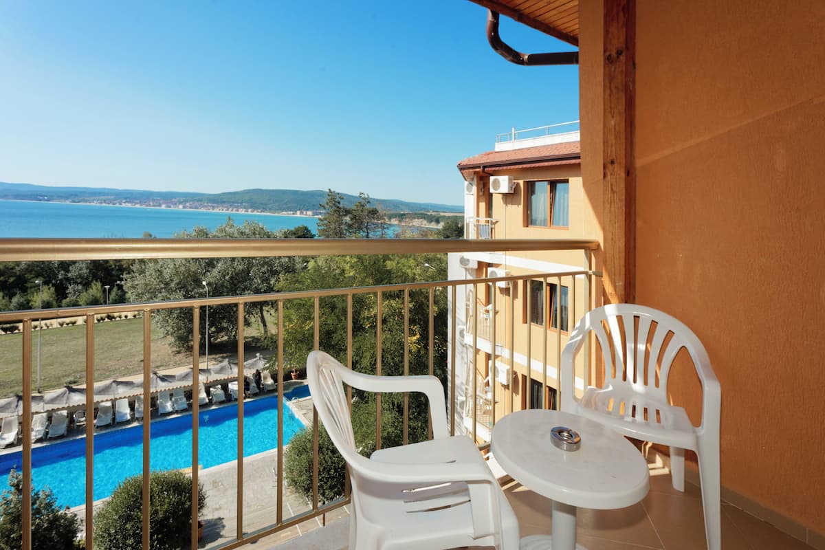 A balcony with a view of a swimming pool and the sea, white plastic chairs, and a small table.
