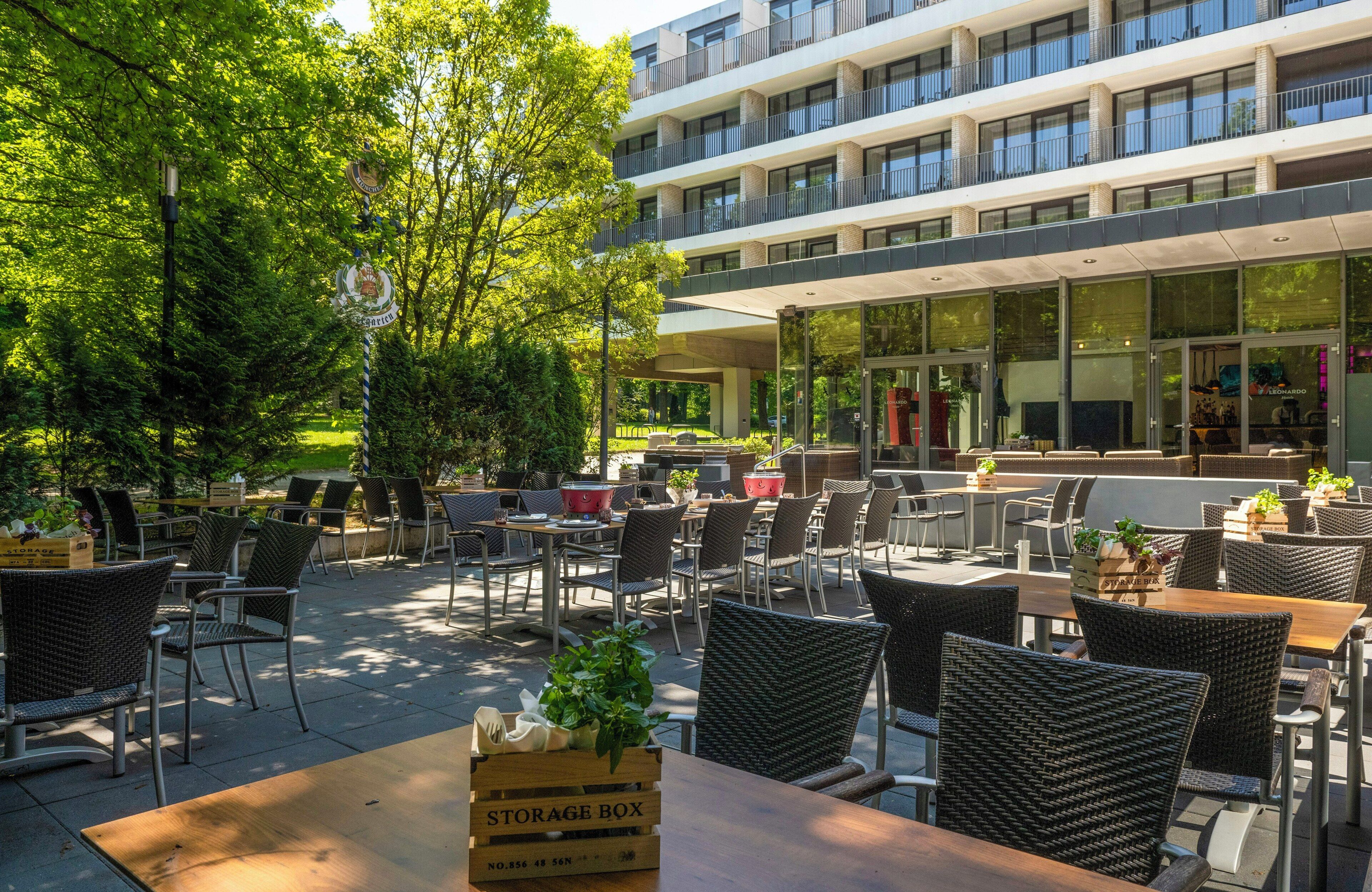 An outdoor cafe with wicker chairs and tables, a storage box, and a building with balconies in the background.
