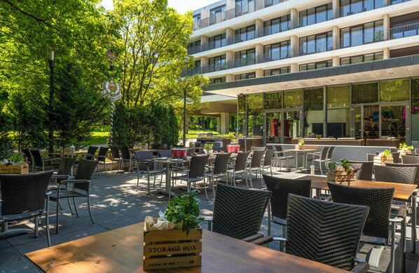 An outdoor cafe with wicker chairs and tables, a storage box, and a building with balconies in the background.