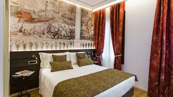 A hotel room with a large mural on the wall, a bed with white linens and patterned pillows, a nightstand with a lamp, and red curtains.