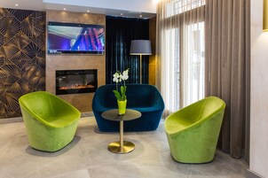 Un área de descanso moderna con un sofá azul, dos sillones verdes, una mesita redonda, una pantalla de televisión y una chimenea.