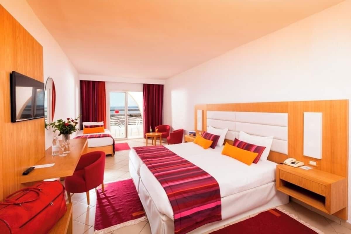 Une chambre d’hôtel avec un grand lit, un bureau avec une chaise, une petite table et une valise rouge.