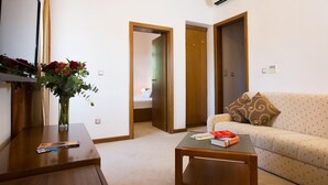 Habitación de hotel con una mesa de madera, un sofá, un jarrón con flores y una cama al fondo.