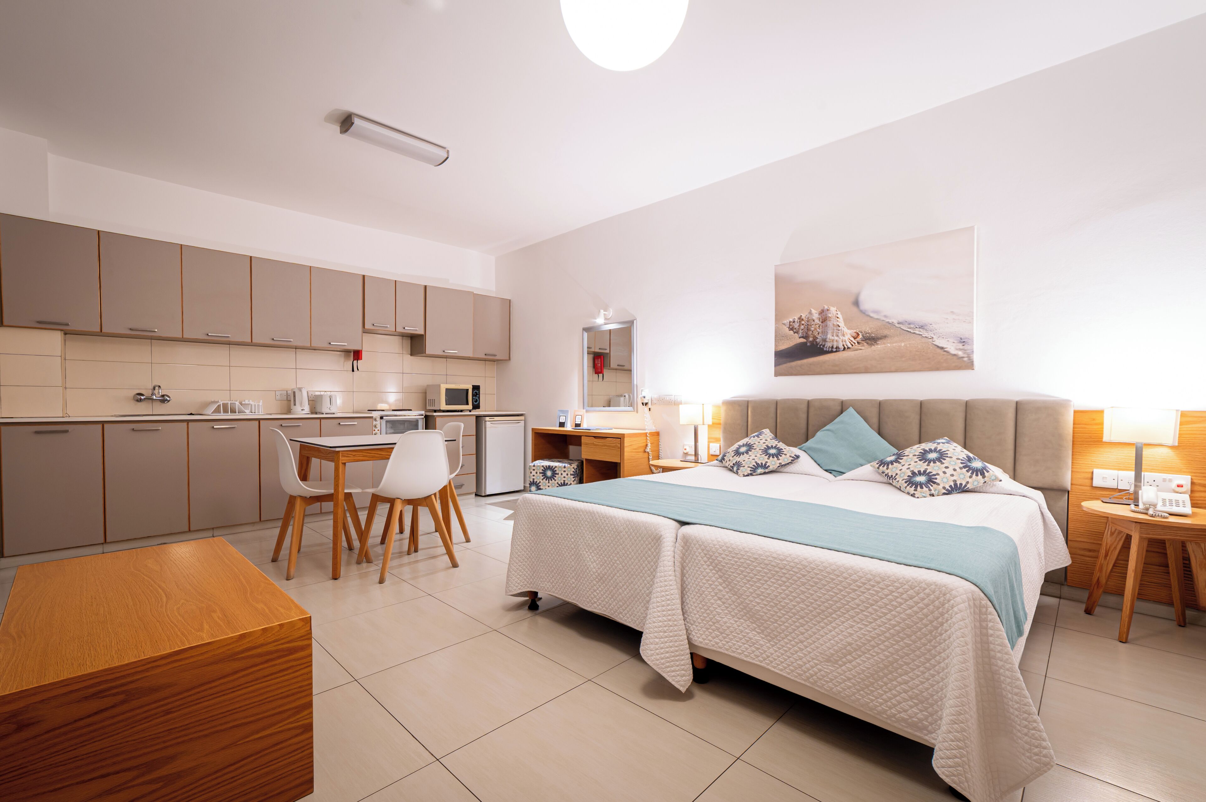 A modern hotel room with a bed, bedside tables, a desk, and a kitchenette.