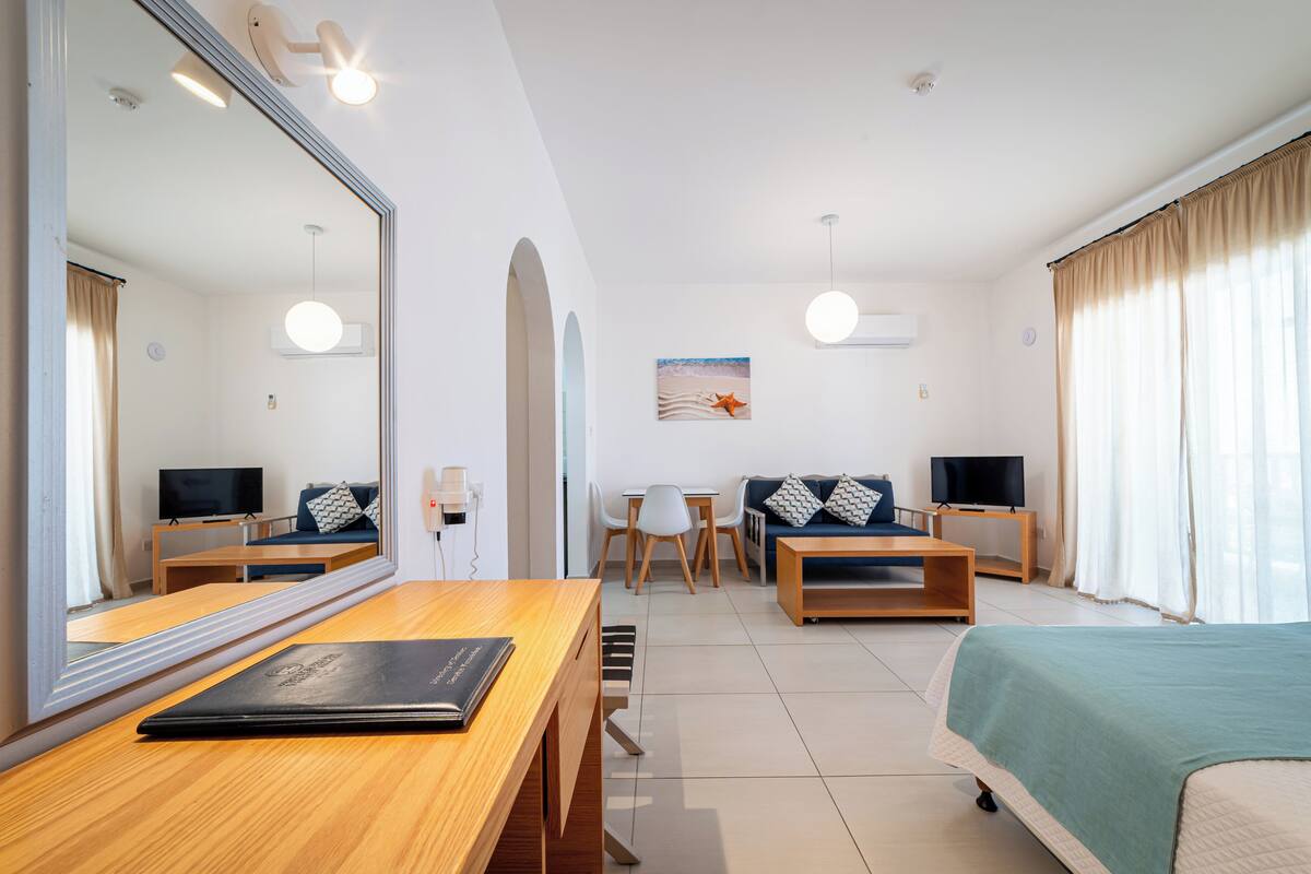 Une chambre d’hôtel avec un grand miroir, un bureau en bois, un lit recouvert d’une couverture verte, une télévision et un tableau accroché au mur.