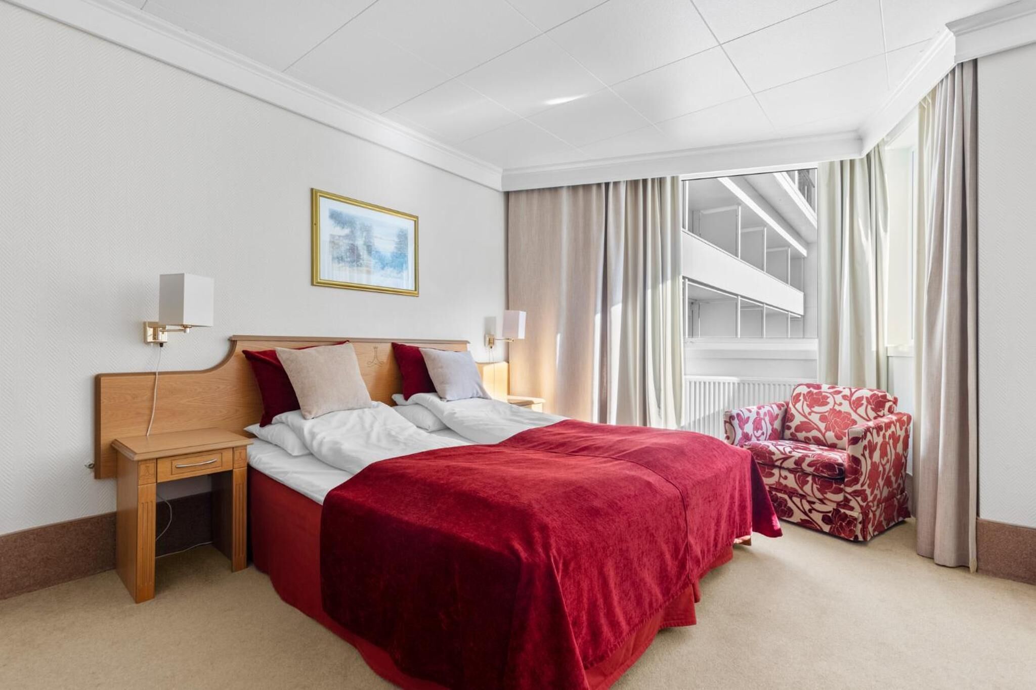 A hotel room with a large bed, a wooden nightstand, a chair, a painting on the wall, and a view of a balcony.