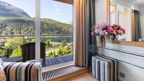 En balkong med utsikt över en sjö och berg, ett glasbord med en vas med blommor och en randig bänk.
