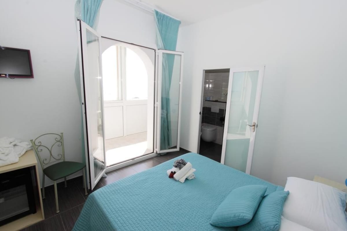 A bedroom with a bed, a chair, a TV, and a sliding door leading to a balcony.