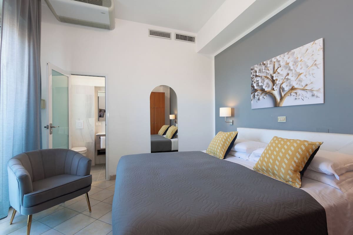 A hotel room with a large bed, a sofa, a painting on the wall, and a bathroom visible through an archway.