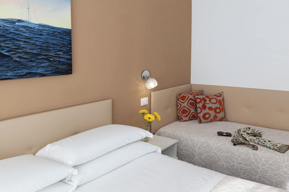 A hotel room with two beds, a painting of the sea, a wall-mounted light, and a vase with flowers.