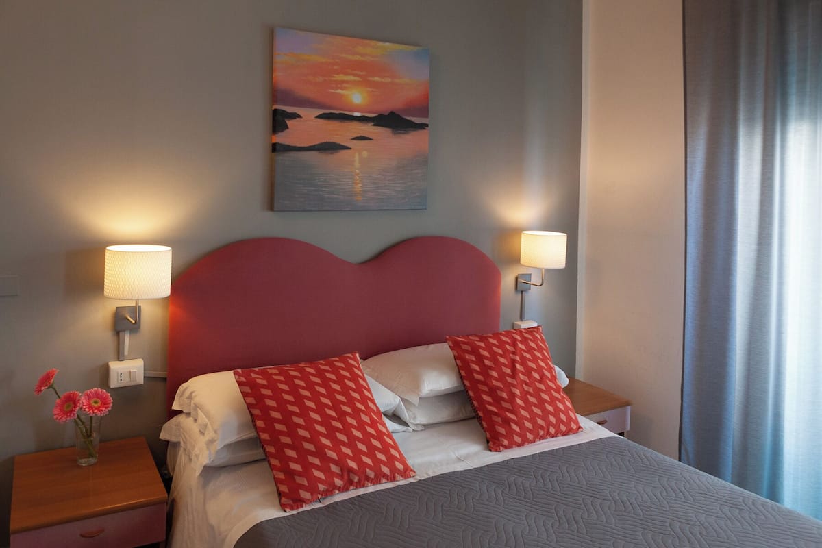 A hotel room with a bed, two bedside lamps, a painting of a sunset, and a window with curtains.