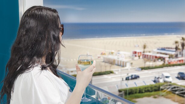 A person holding a glass of white wine, overlooking a beach with buildings and palm trees in the background. - MoMo Seaview Hotel Rimini (Rimini)