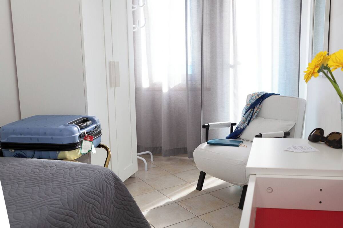 A hotel room with a bed, a desk with a vase of flowers, a chair, and a suitcase.