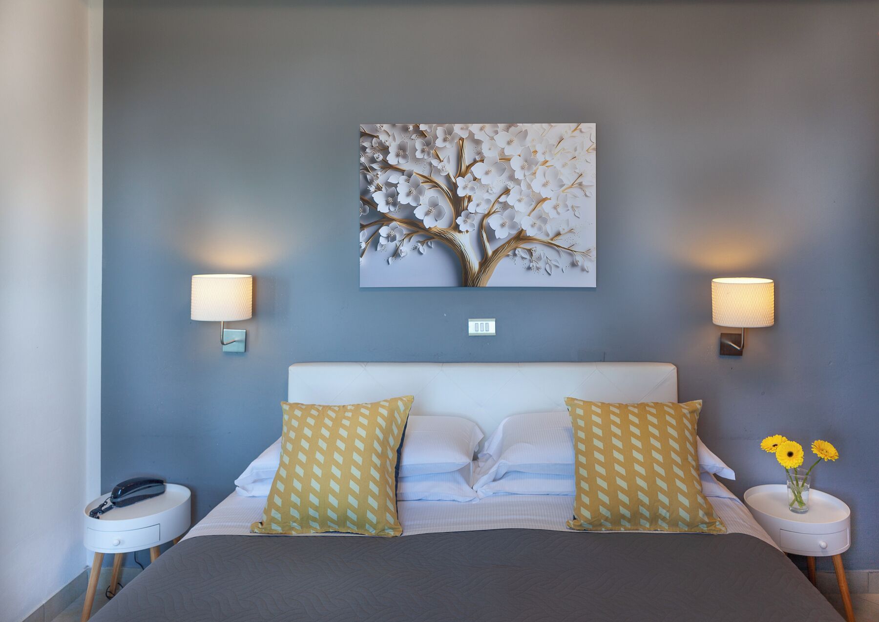 A bedroom with a bed, two bedside tables, a wall-mounted lamp, and a floral artwork.