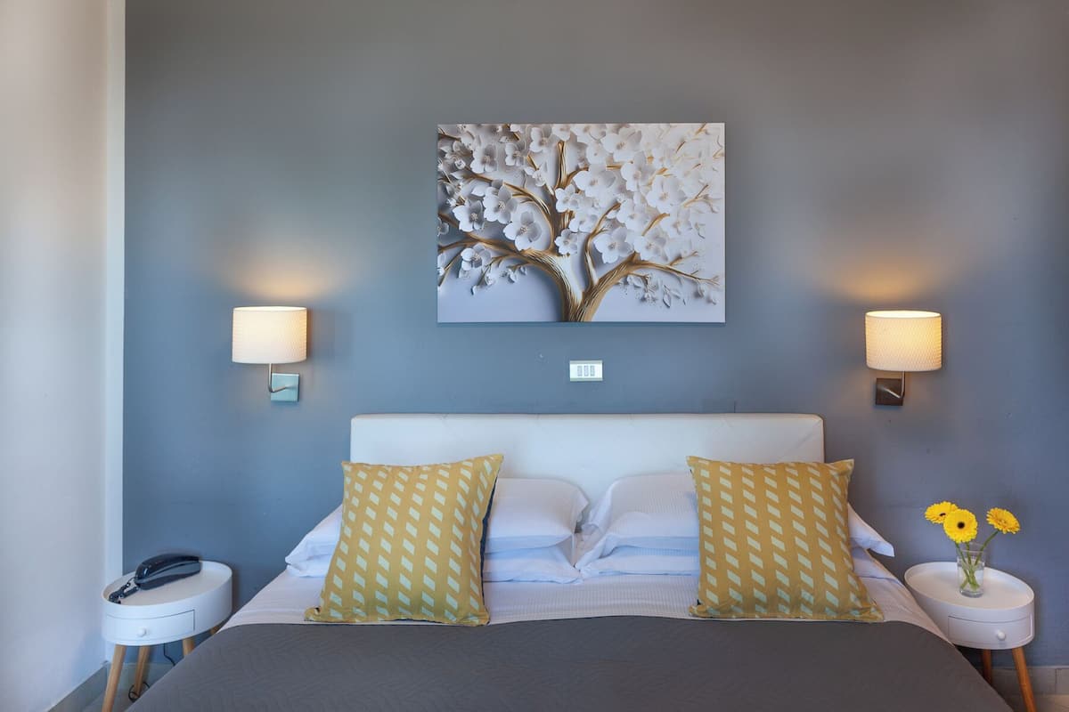 A bedroom with a bed, two bedside tables, a wall-mounted lamp, and a floral artwork.
