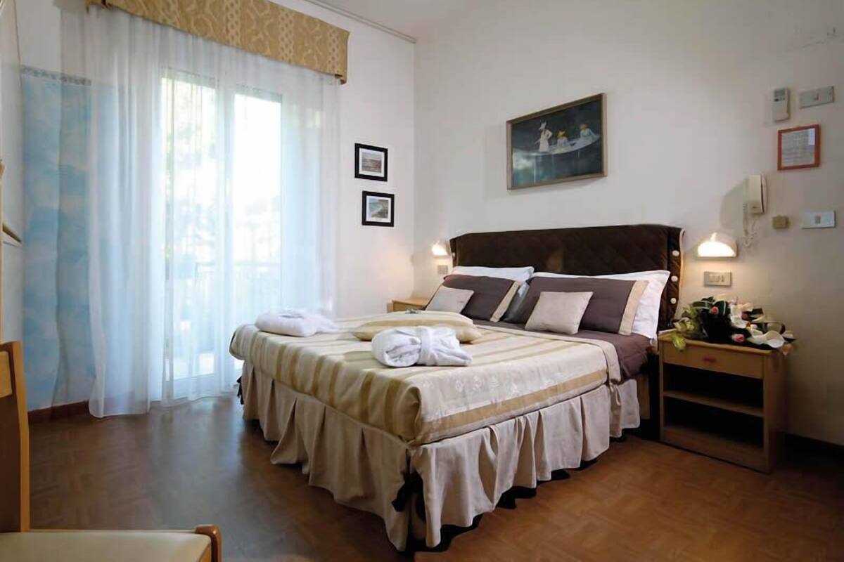 A double bed with a headboard, two bedside tables, a nightstand, and a window with sheer curtains.