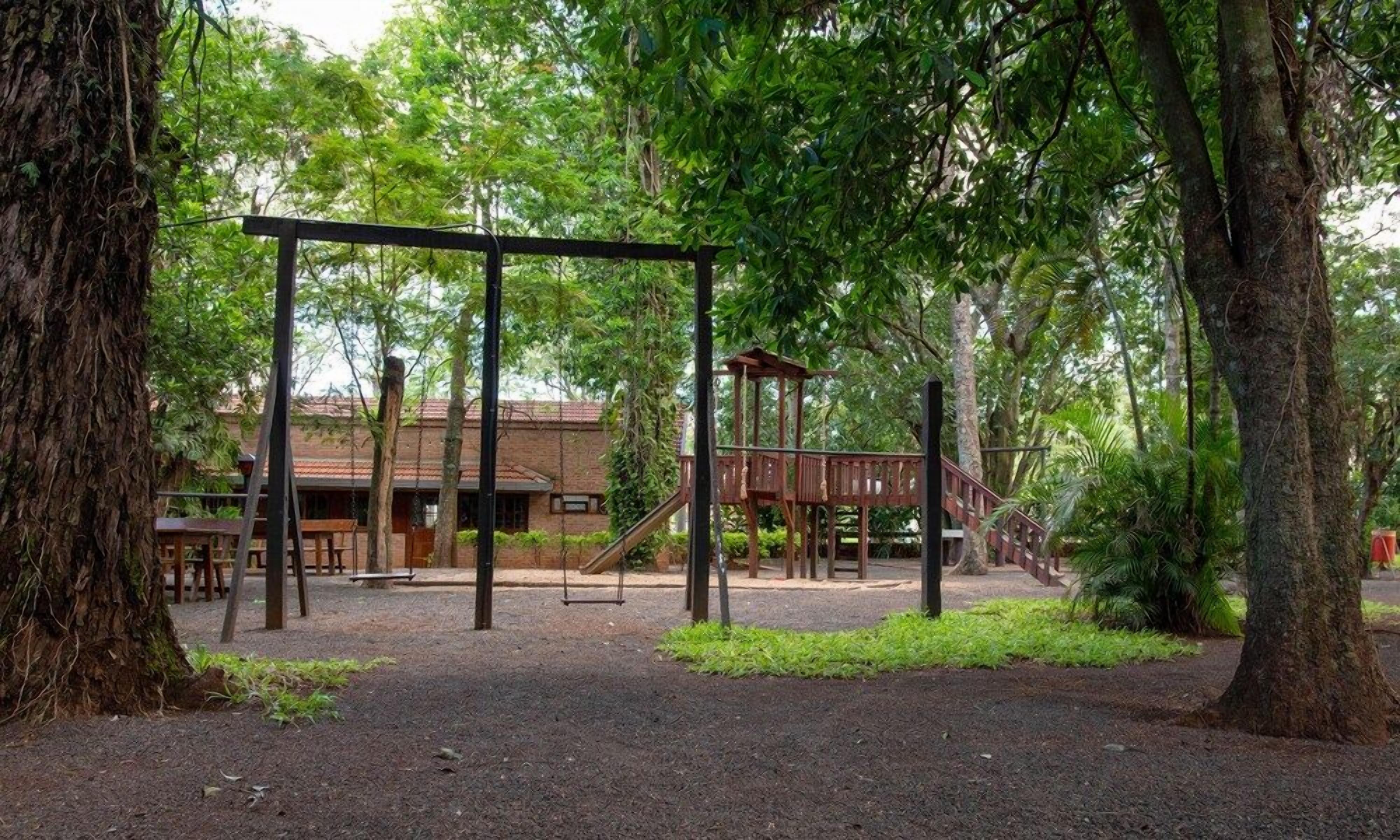 A playground with swings and a slide, surrounded by trees and a building in the background.