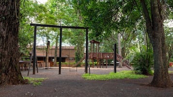 A playground with swings and a slide, surrounded by trees and a building in the background.