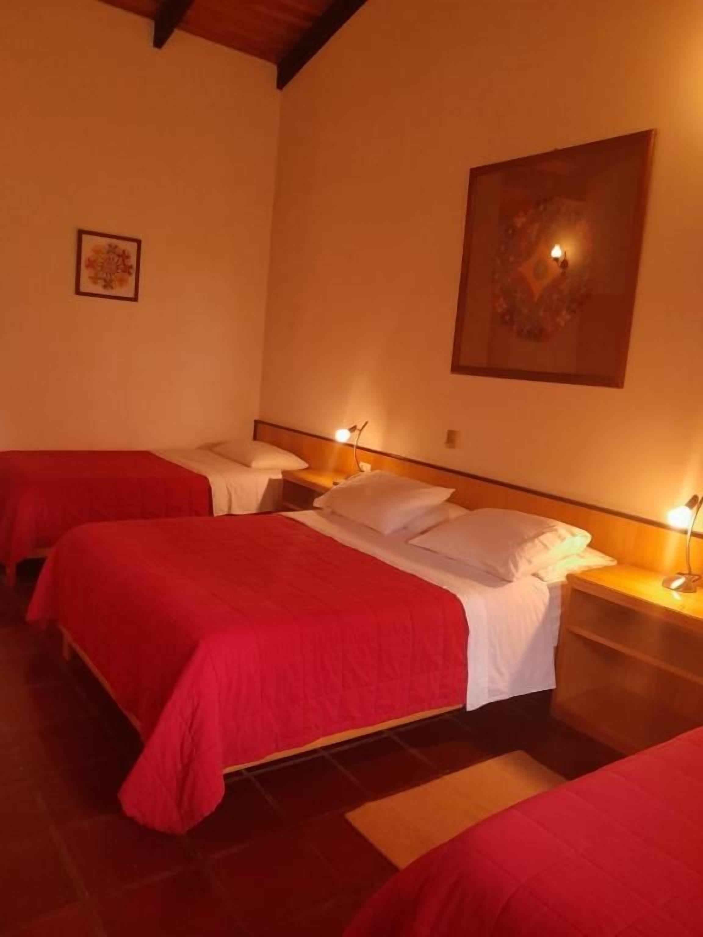 A hotel room with two beds, each with a red bedspread, a nightstand with a lamp, and a painting on the wall.