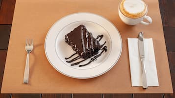 Een plak chocoladecake met chocoladesaus op een wit bord, een kop koffie, een vork en een mes op een bruine placemat.