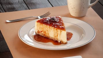 A slice of cheesecake on a plate with a fork and knife, a cup of coffee, and a napkin on a wooden table.