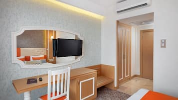 Ett hotellrum med ett skrivbord i trä, en stol, en tv, en garderob och en säng med ett orangefärgat överkast.