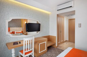 Habitación de hotel con escritorio de madera, una silla, televisión, armario y cama con colcha naranja.