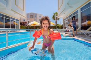 Zona de piscina con agua azul cristalina, un niño en traje de baño con flotadores y un edificio con grandes ventanales y sombrillas al fondo.