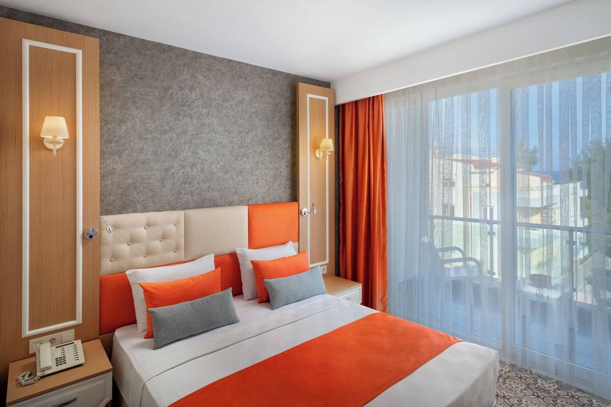 A hotel room with a bed, a telephone, a window with curtains, and a view of the outside.