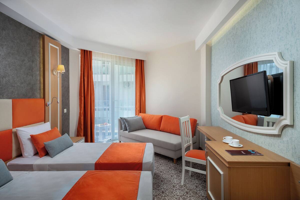 A hotel room with two beds, a sofa, a desk, a chair, a TV, and a window with curtains.