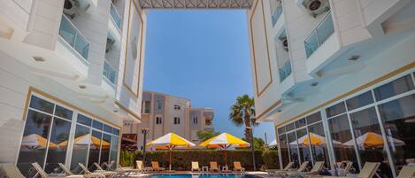 A swimming pool with sun loungers and umbrellas, surrounded by a modern building with large windows and a clear sky.