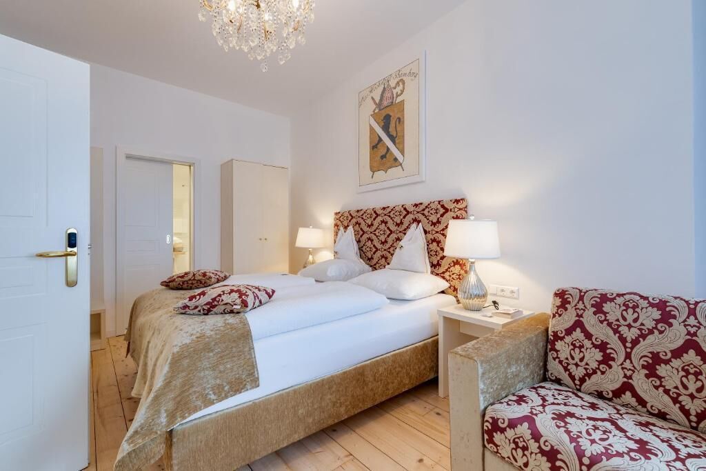 Une chambre d’hôtel avec un lit, des lampes de chevet, un fauteuil à motifs et un lustre.