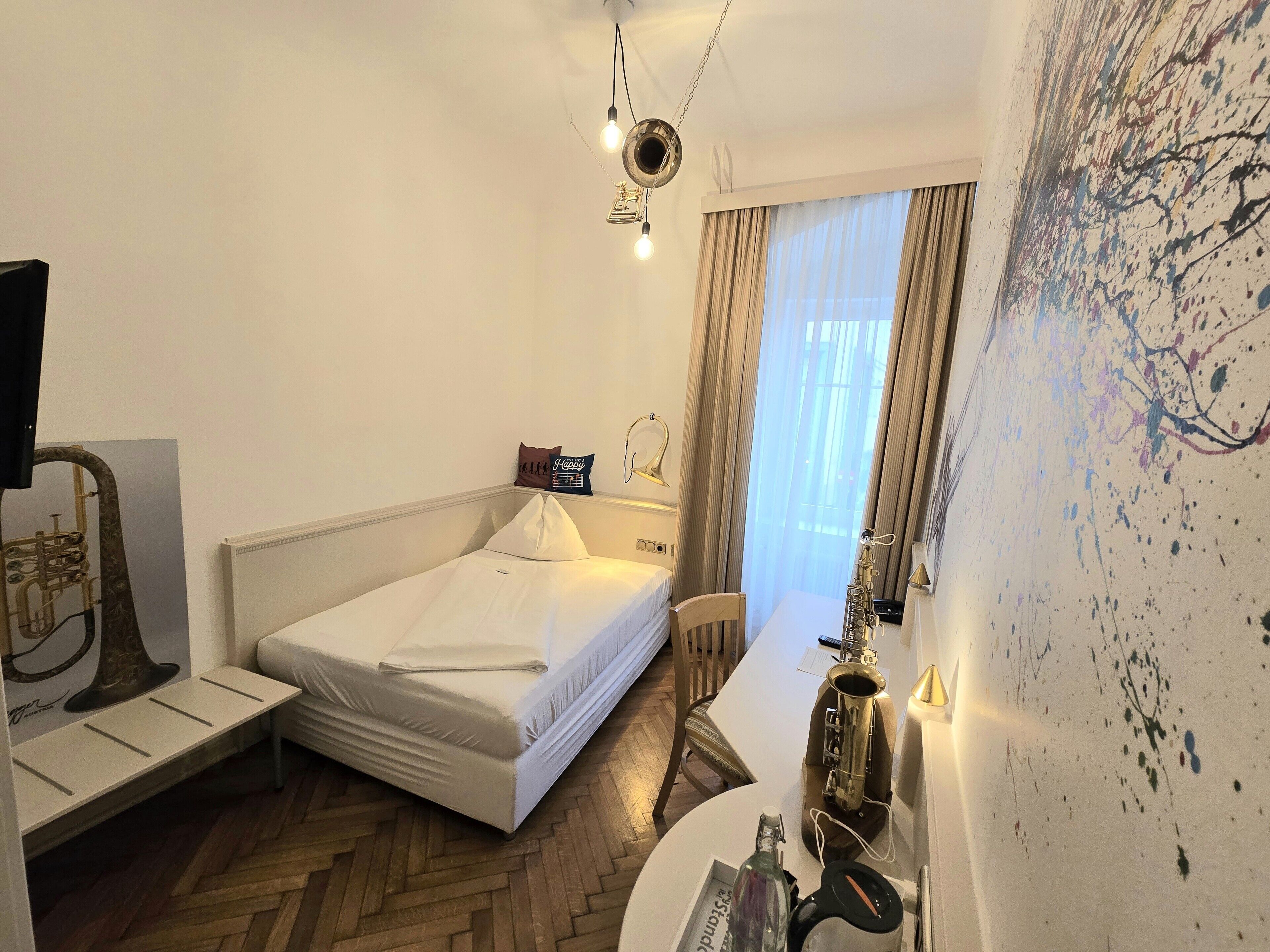 Habitación de hotel con cama, escritorio, silla, un saxofón y un cuadro abstracto en la pared.