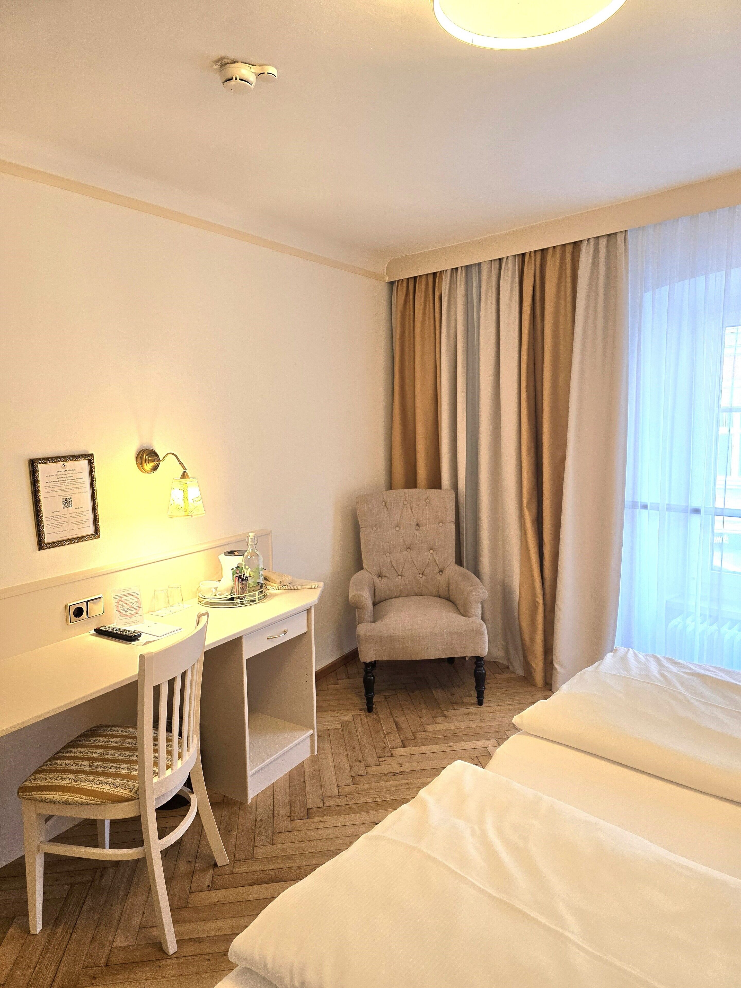 Quarto de hotel com cama, escrivaninha, cadeira, luminária e janela com cortinas.