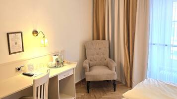 A hotel room with a bed, a desk with a chair, a lamp, and a window with curtains.