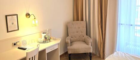 Quarto de hotel com cama, escrivaninha, cadeira, luminária e janela com cortinas.