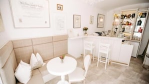 Une pièce moderne et bien éclairée, avec un canapé beige, une table ronde blanche et des chaises blanches. On y trouve également un coin bar avec des étagères remplies de bouteilles et de verres.